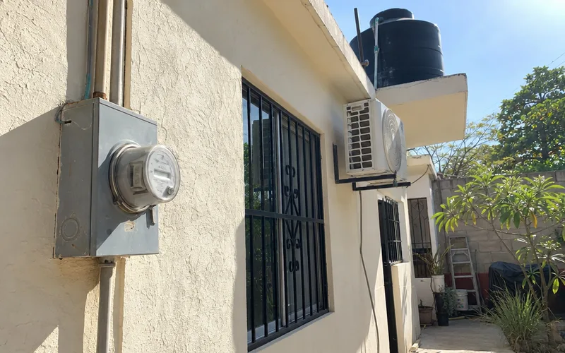 Electric meter and wall-mounted air conditioner outside a stucco house in Mexico with a rooftop water tank