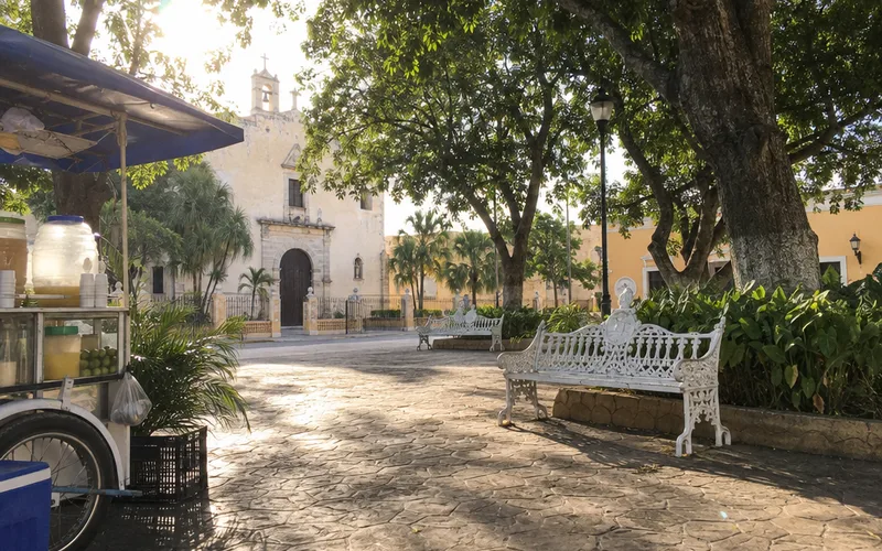 Shaded benches under large trees in a quiet plaza in Mérida with a church in the background